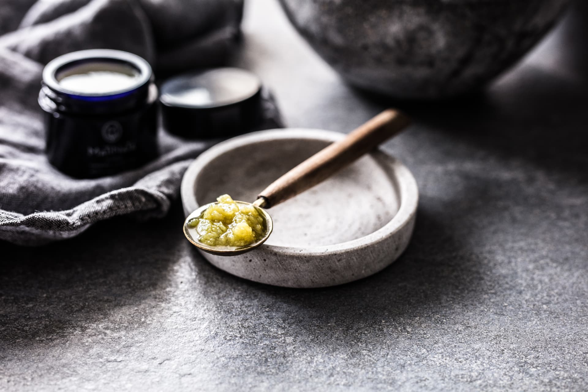 The making of Matinata — a spoon lifting freshly pressed botanical balm above a stone bowl, beside a dark blue jar and folded linen on slate